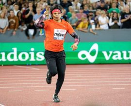 Image of Neeraj Chopra, a muscular Indian athlete, holding a javelin in his throwing position. He is wearing an athletic uniform and has a determined expression on his face.