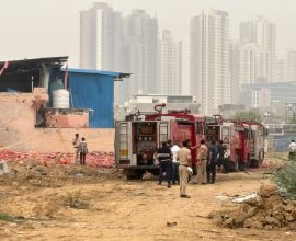 Gurgaon Factory Blast - Firefighters and authorities inspect the aftermath of an explosion at a factory site in Gurgaon, with high-rise buildings in the background.