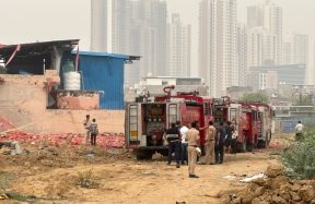 Gurgaon Factory Blast - Firefighters and authorities inspect the aftermath of an explosion at a factory site in Gurgaon, with high-rise buildings in the background.