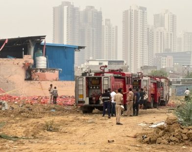 Gurgaon Factory Blast - Firefighters and authorities inspect the aftermath of an explosion at a factory site in Gurgaon, with high-rise buildings in the background.