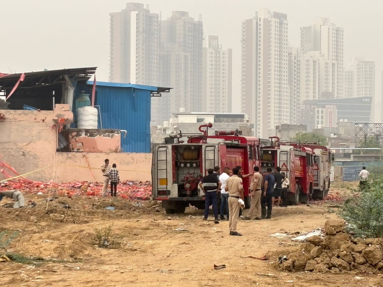 Gurgaon Factory Blast - Firefighters and authorities inspect the aftermath of an explosion at a factory site in Gurgaon, with high-rise buildings in the background.