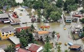 A flooded street scene in Sri Lanka. Floodwaters rise above cars and houses, some submerged entirely. People wade through the water, some carrying belongings.