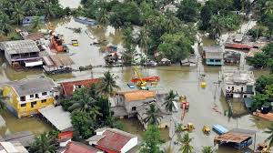 A flooded street scene in Sri Lanka. Floodwaters rise above cars and houses, some submerged entirely. People wade through the water, some carrying belongings.