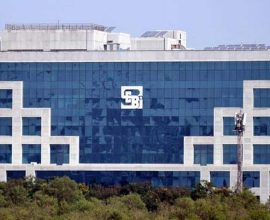 large glass building with the logo of the Securities and Exchange Board of India (SEBI) in the center. There are trees in the background.