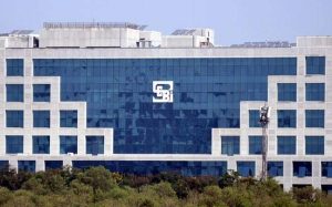 large glass building with the logo of the Securities and Exchange Board of India (SEBI) in the center. There are trees in the background.